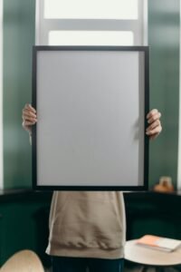 Anonymous person holds a blank vertical picture frame indoors against a window.